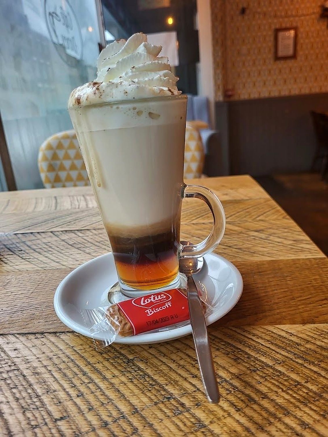 A tall layered hot drink topped with whipped cream, a Lotus Biscoff biscuit on the saucer