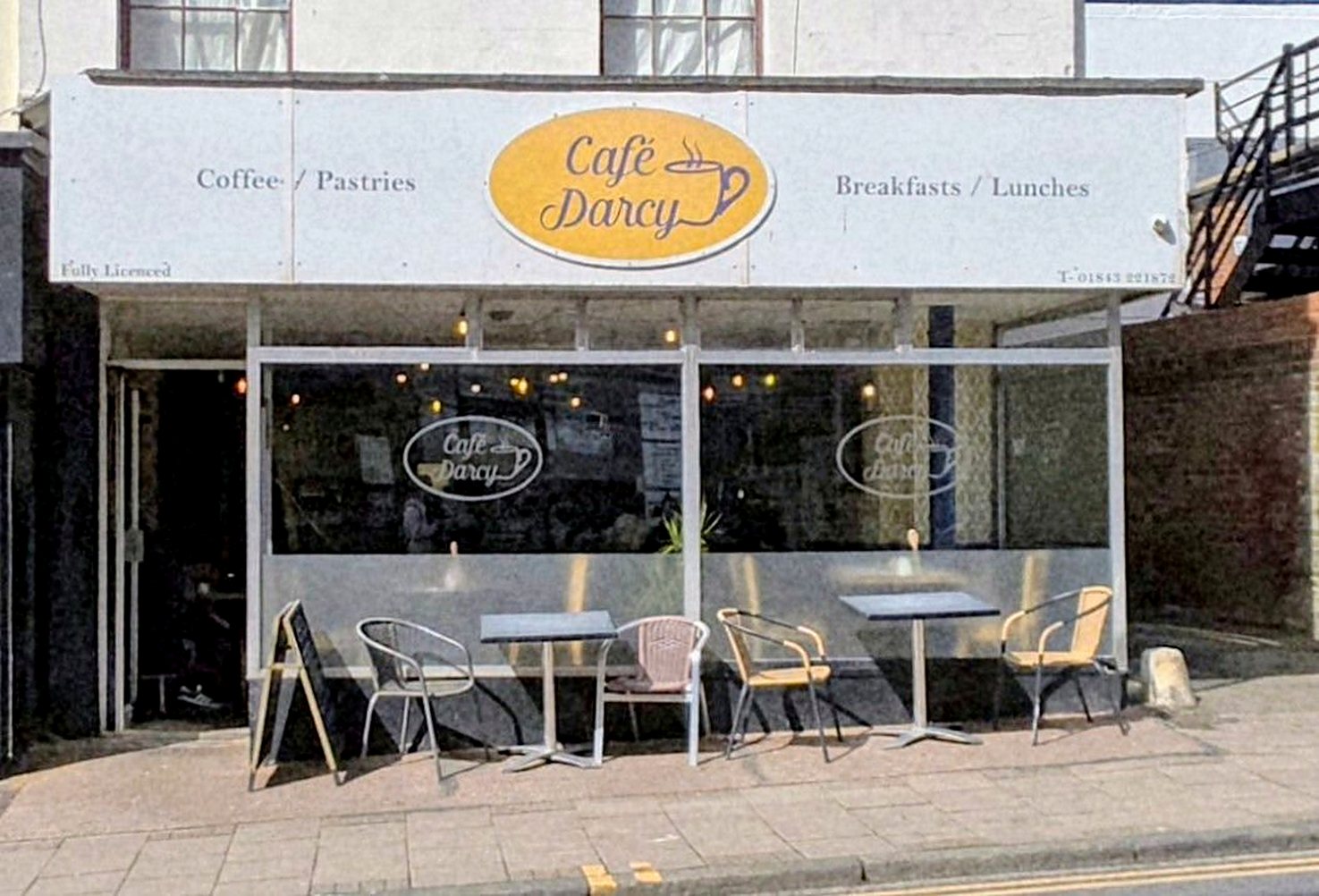 Café Darcy on Marine Gardens: white fascia with a yellow oval sign, big windows and bistro chairs on the pavement